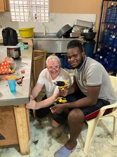 An elderly woman and a young man sitting together in a kitchen, smiling, with the woman holding a yellow power drill.