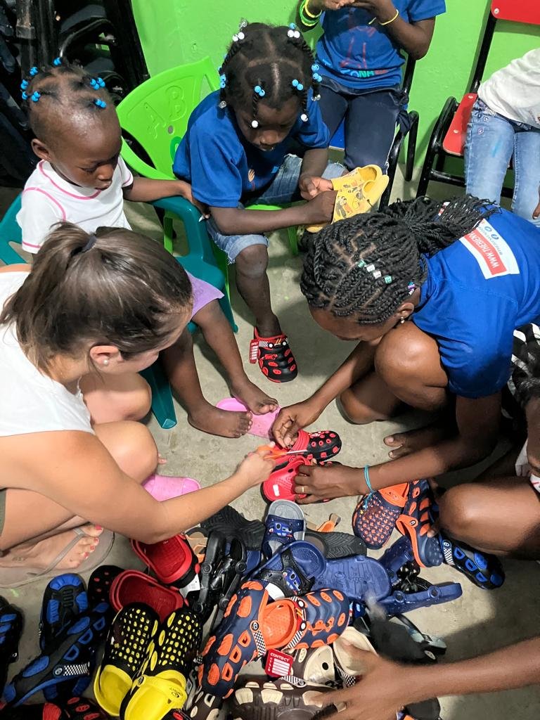 Children sitting in a circle on the floor, trying on pairs of shoes with the help of an adult. There are stacks of shoes on the ground in front of them.