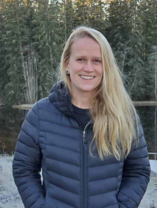 A smiling woman with long blonde hair standing outdoors near a body of water and trees.