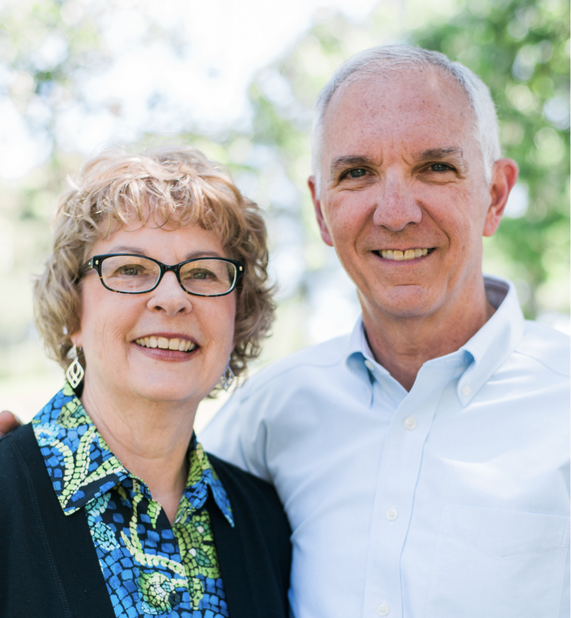 A smiling elderly woman with glasses and curly hair standing next to a smiling elderly man with short white hair, both outdoors with blurred green trees in the background.