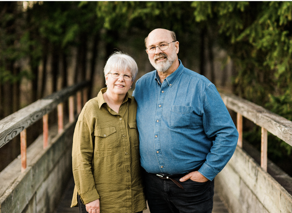 An elderly couple standing on a wooden bridge surrounded by greenery, smiling at the camera.