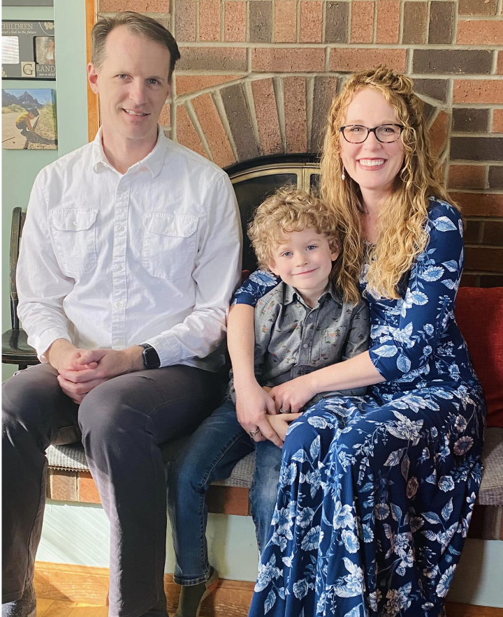 A family of three sitting on a bench in front of a brick fireplace. The man on the left wears a white shirt, the woman on the right wears glasses and a blue floral dress, and a young boy sits between them with curly blonde hair, wearing a gray shirt.