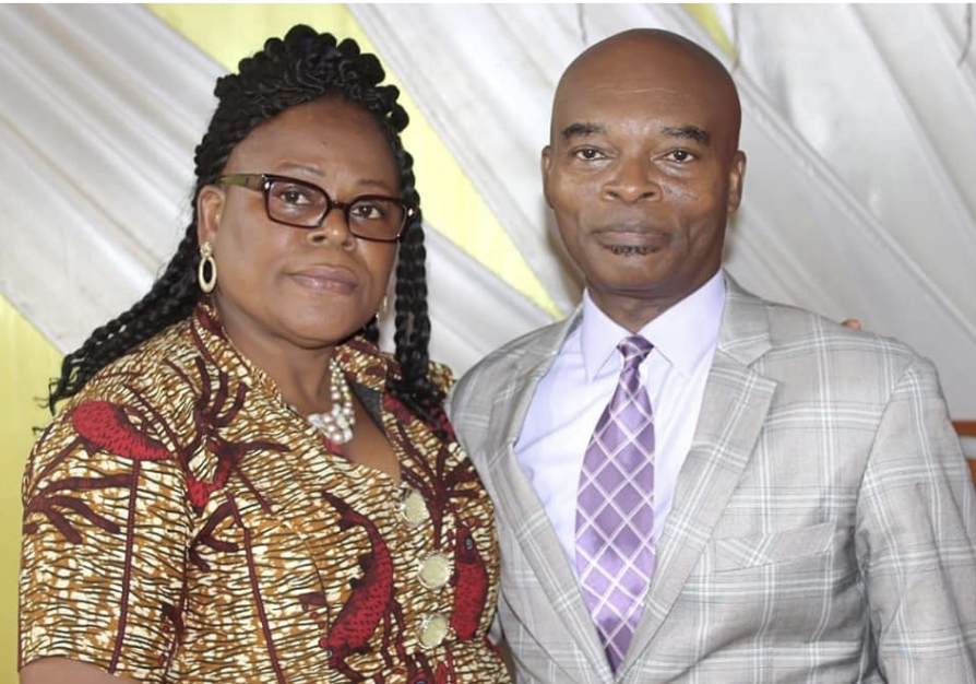 A woman and a man in formal attire posing together indoors, with a light-colored draped background.