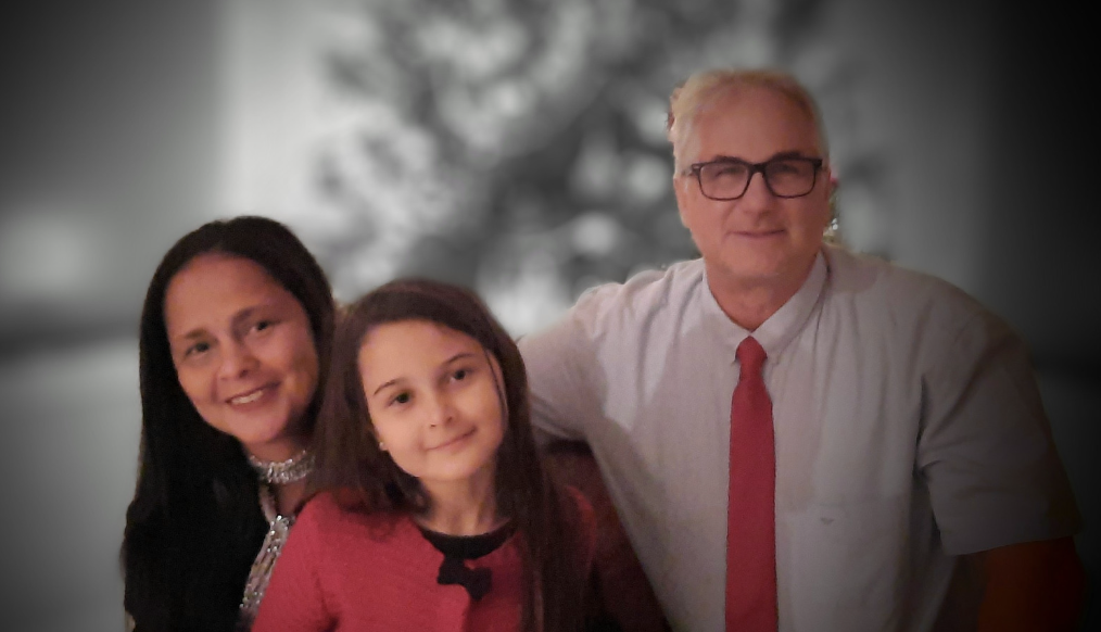 A man with glasses, white hair, and a white shirt with a red tie, standing with two young girls, one with dark hair and a black top, and the other with brown hair and a red top, all smiling