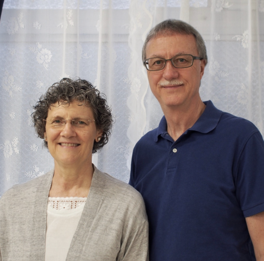 A smiling elderly woman with curly gray hair and glasses standing next to an elderly man with short gray hair, glasses, and a blue polo shirt, in front of a white lace curtain background.