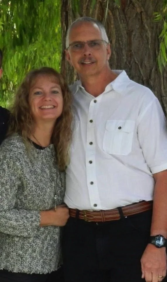 A smiling middle-aged woman with curly hair and a middle-aged man with glasses, standing outdoors in front of a tree with green foliage.