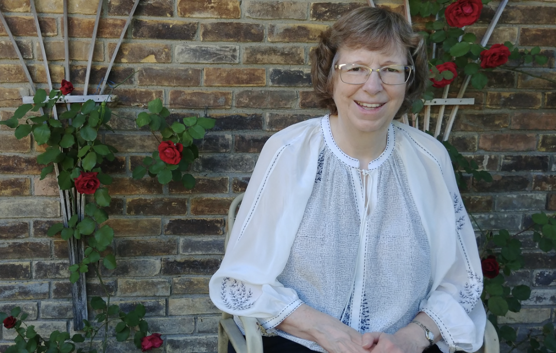 A woman with glasses and short curly hair, seated outdoors in front of a brick wall with red roses on trellises.