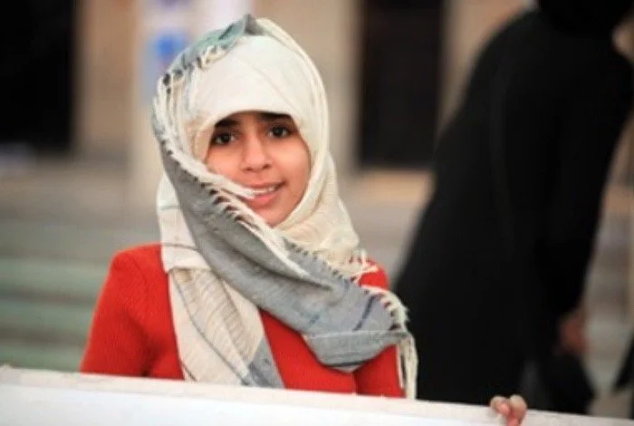 A young girl wearing a white headscarf and a red sweater, smiling at the camera.