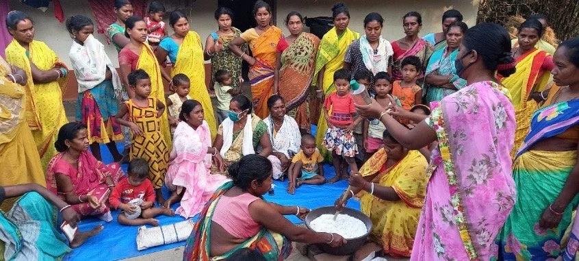 Group of women and children gathered outdoors around a woman demonstrating cooking, with some people sitting on a blue mat.