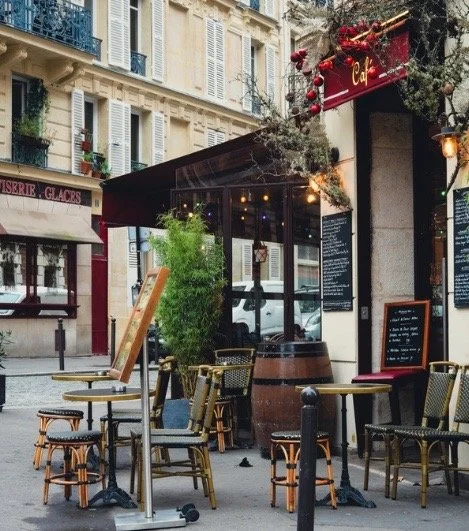 Outdoor seating area of a cafe with several chairs and tables, some with umbrellas, in front of a sidewalk restaurant on a city street, decorated with a sign and string lights.