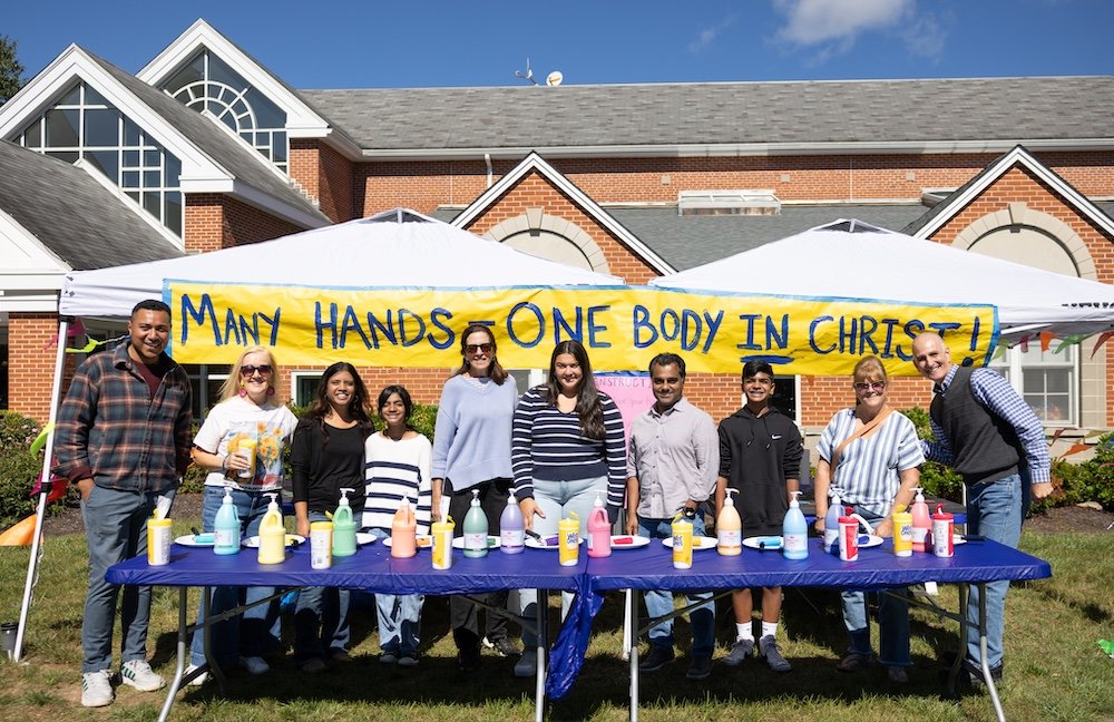 A group of nine people standing behind a table with various bottles of paint, in front of a white tent with a yellow banner that reads 'Many Hands One Body in Christ.' They are outdoors in front of a house with a brick facade and large windows.