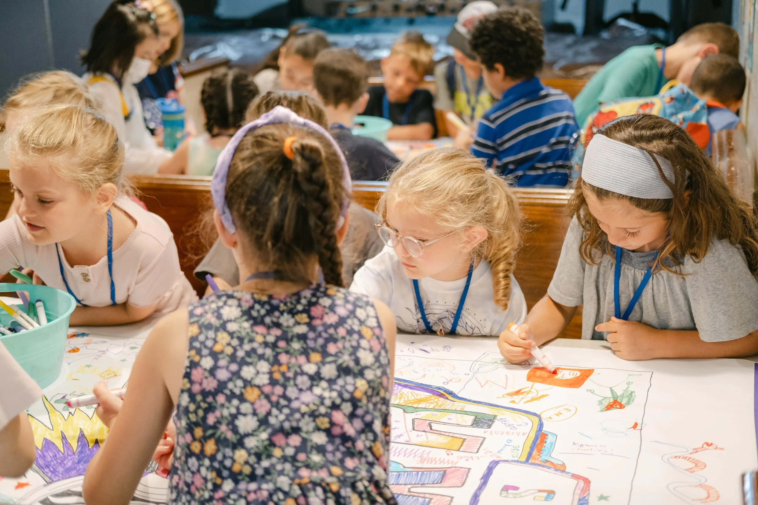 Children sitting at a table coloring and drawing on a large sheet of paper with markers, with more children visible in the background.