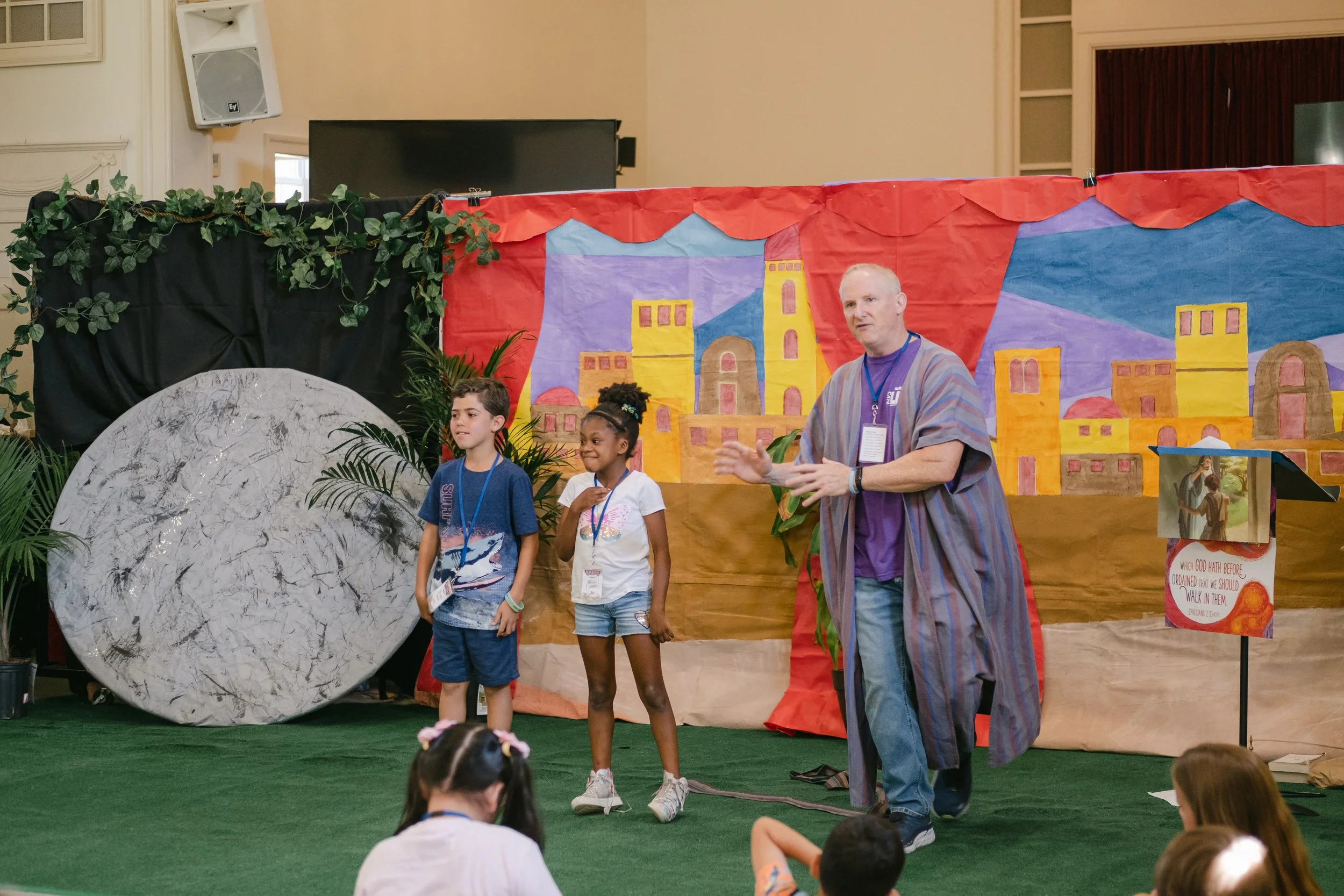 A man in a striped robe and purple shirt is speaking to an audience of children, with two children standing on stage near him, in front of a colorful backdrop depicting a cityscape with tall buildings and a castle, surrounded by fake plants and a lar