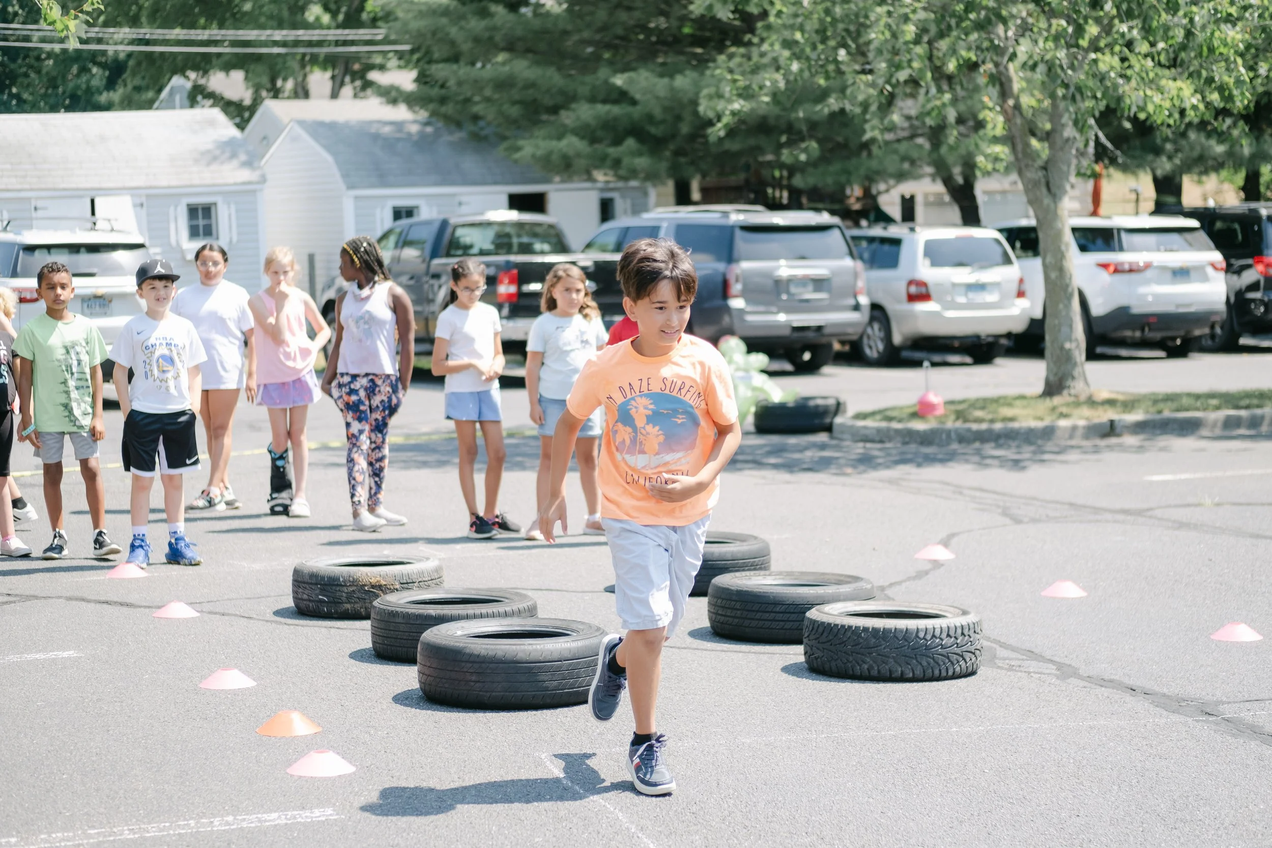 A group of children participating in an outdoor activity involving running and tires, with cones marking the course, on a paved area near parked cars and trees.
