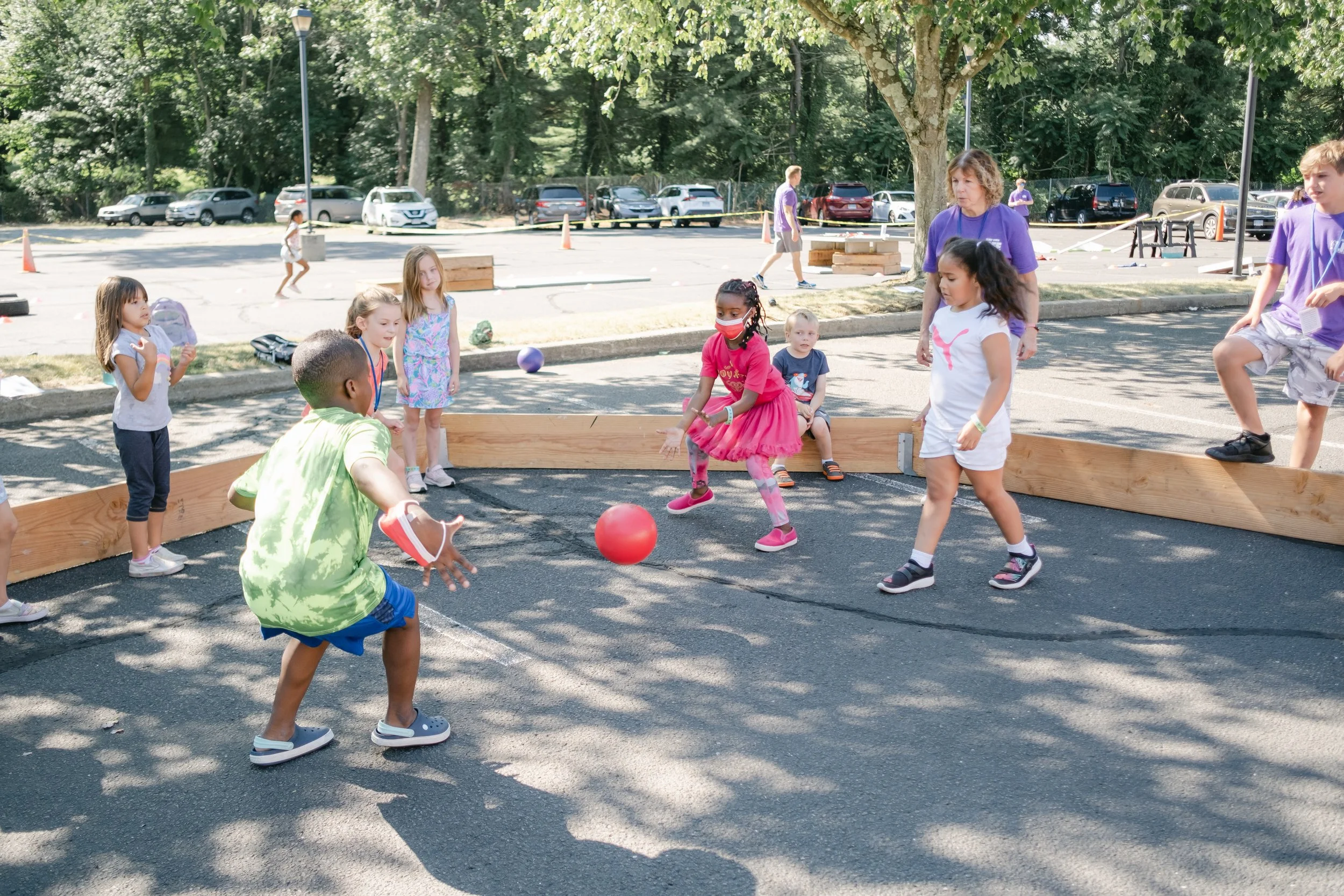 Children playing in a sandbox with a ball, supervised by adults, outdoors on a sunny day.