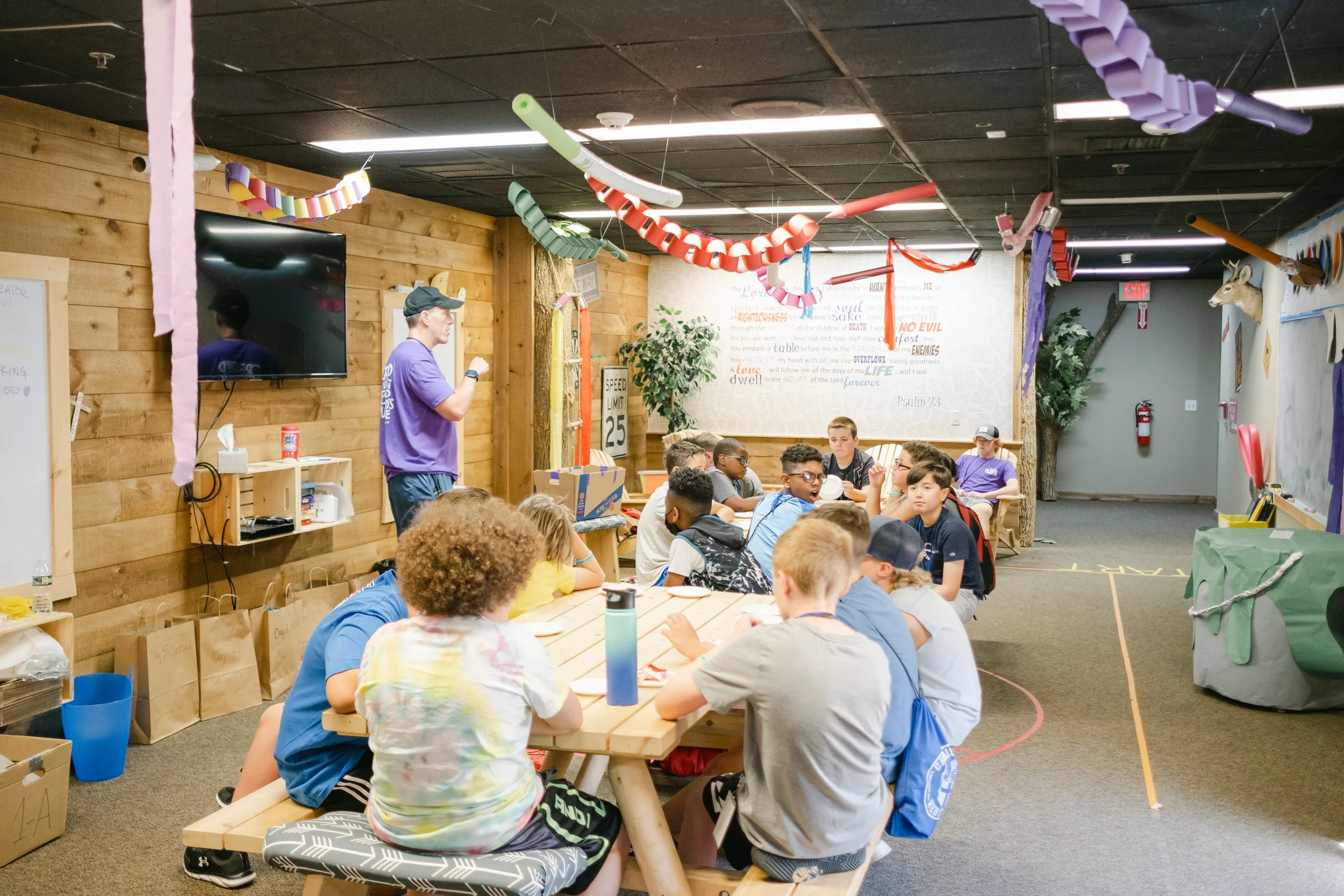 Group of children sitting at a table in a decorated room with streamers, a TV, and wall decorations, during a party or gathering.
