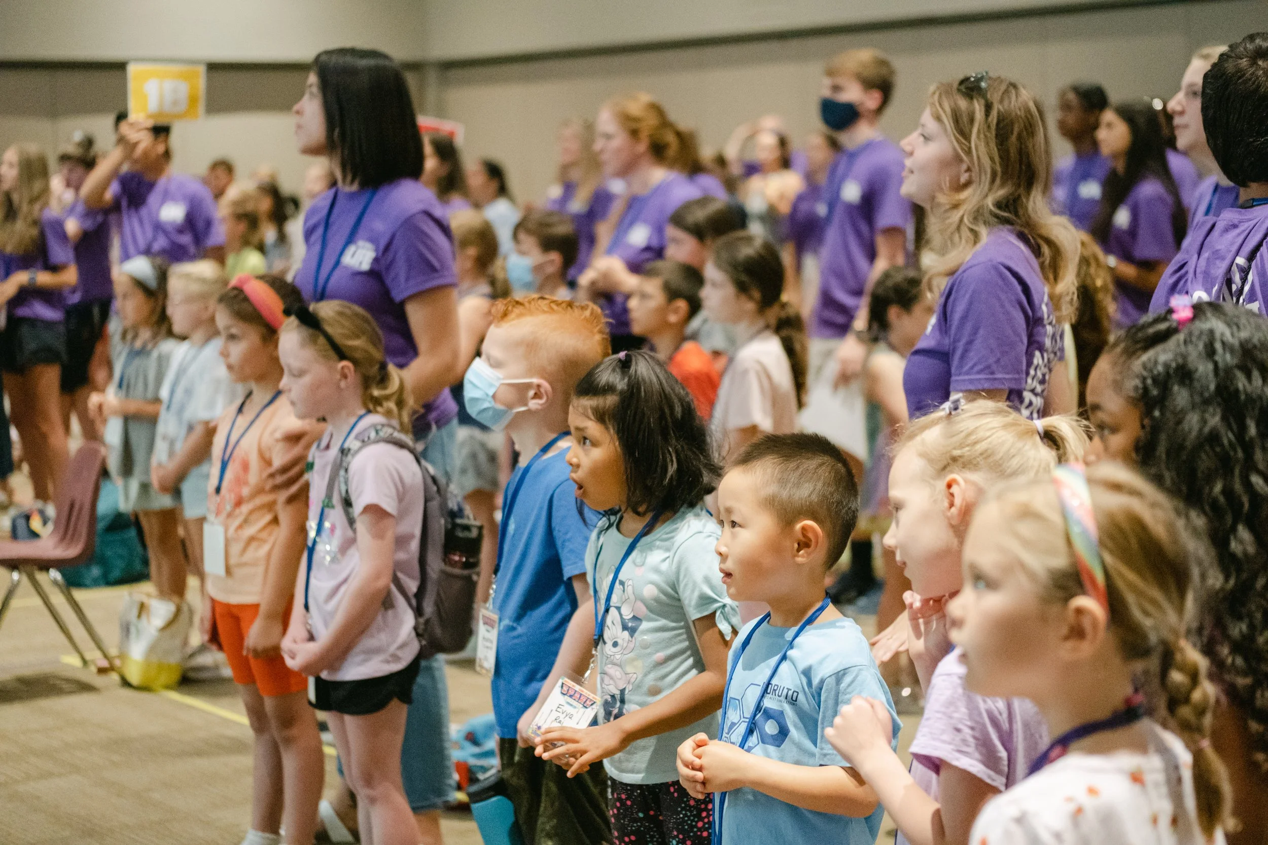 Children and young adults standing in a large indoor room, some wearing face masks, participating in a group activity or event, with many wearing purple shirts and lanyards.