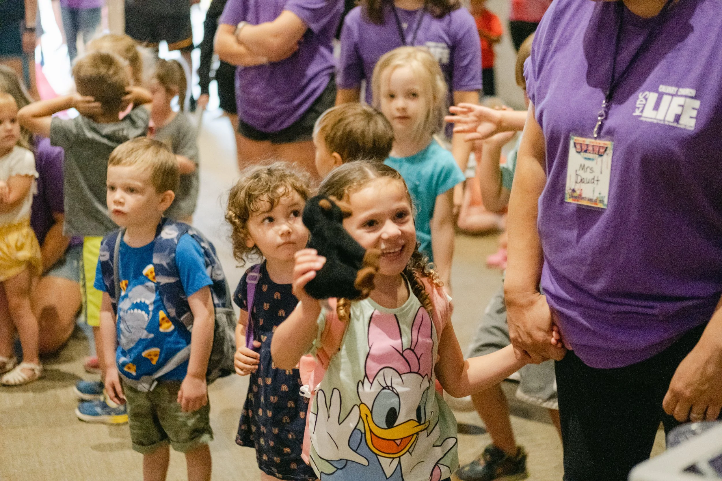 A group of young children standing in line at an indoor event, waiting for an activity. One girl is smiling and holding up a small stuffed animal, while an adult in a purple shirt holds her hand.