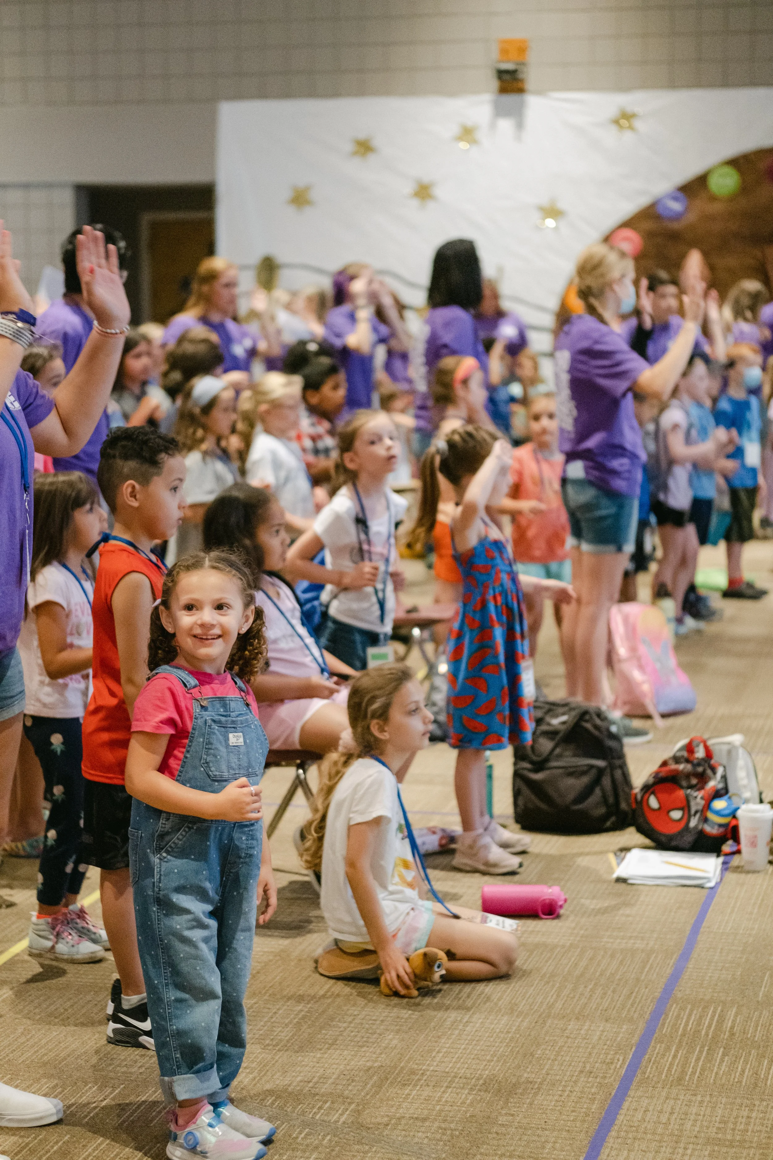 Children and adults standing and sitting, participating in an indoor event or assembly, some raising hands, with a decorated stage or backdrop featuring stars and balloons.