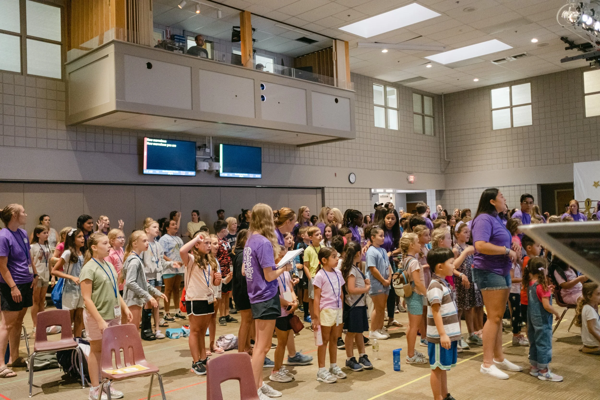 A large group of children and a few adults gathered in a gymnasium or auditorium, standing and facing forward, possibly participating in an event or assembly. Many of the children are wearing lanyards, and some adults are wearing purple shirts.