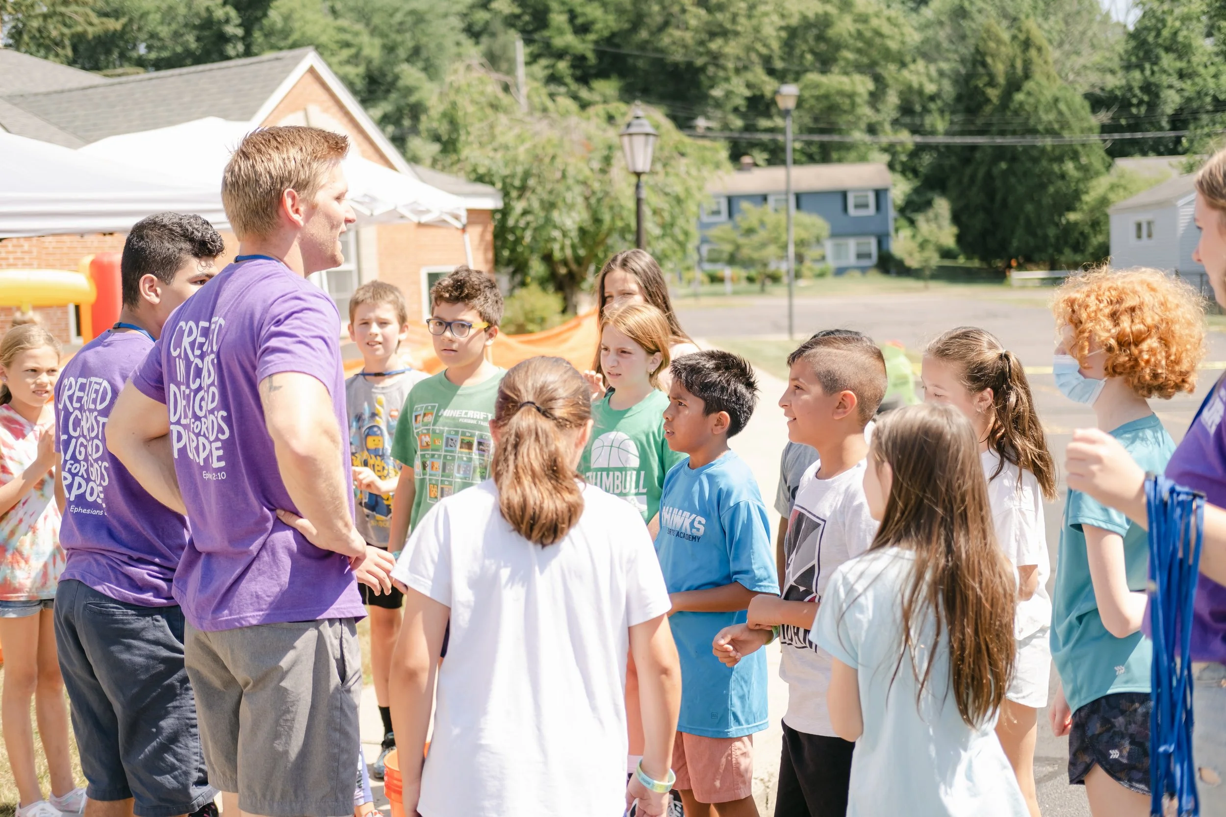 Group of children and camp counselors outdoors at a daytime camp activity, with trees and houses in the background.