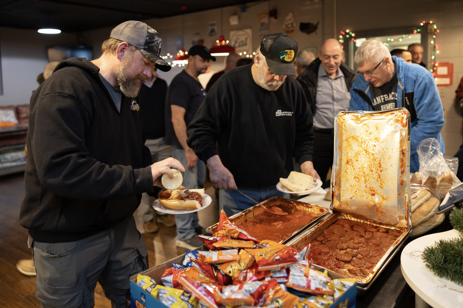 People serving and receiving hot dogs and food at a buffet table during a social gathering in a decorated indoor space.