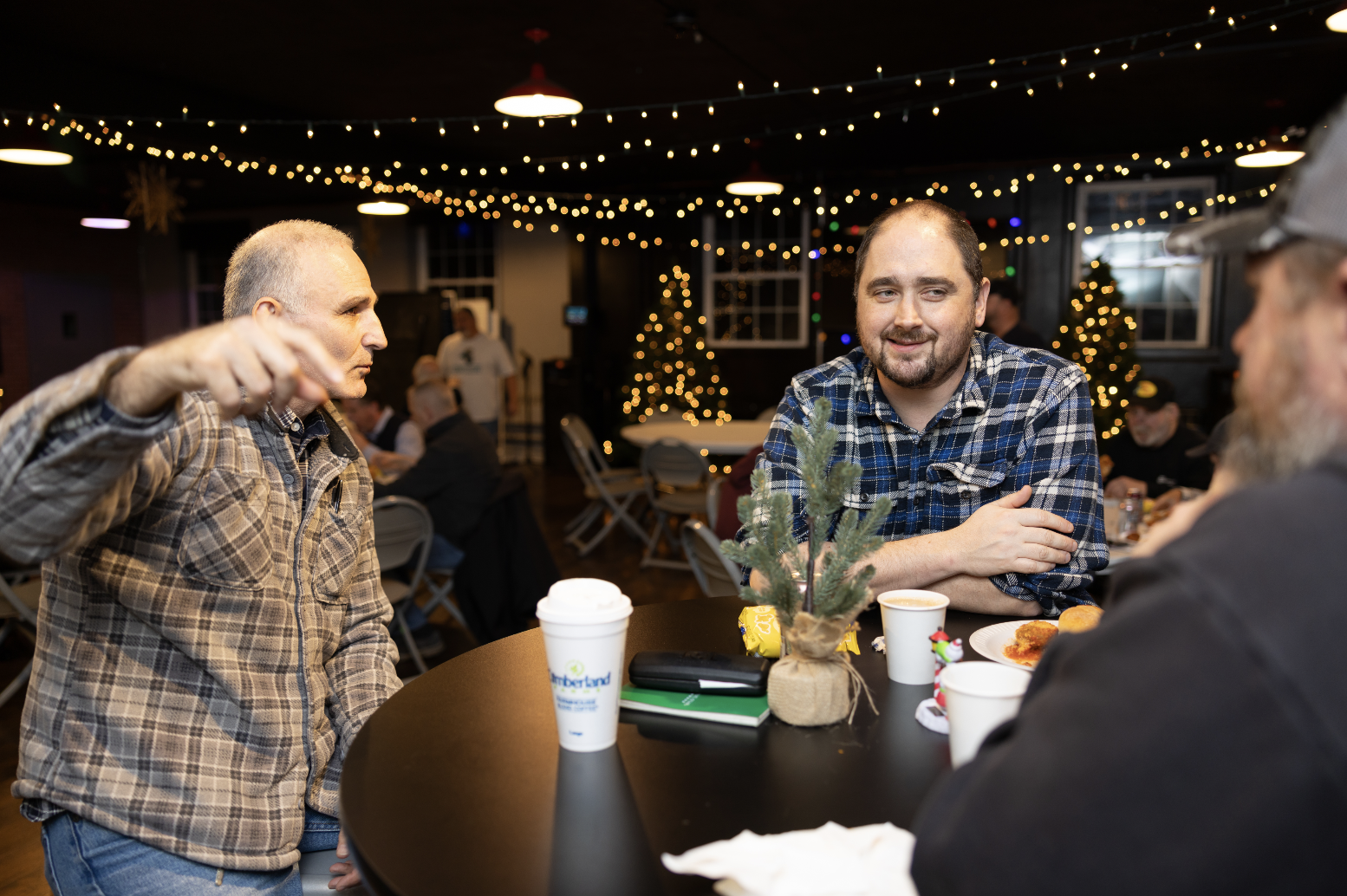 People gathered around a table decorated with a small Christmas tree, with holiday lights and ornaments visible in the background, during a festive holiday gathering.