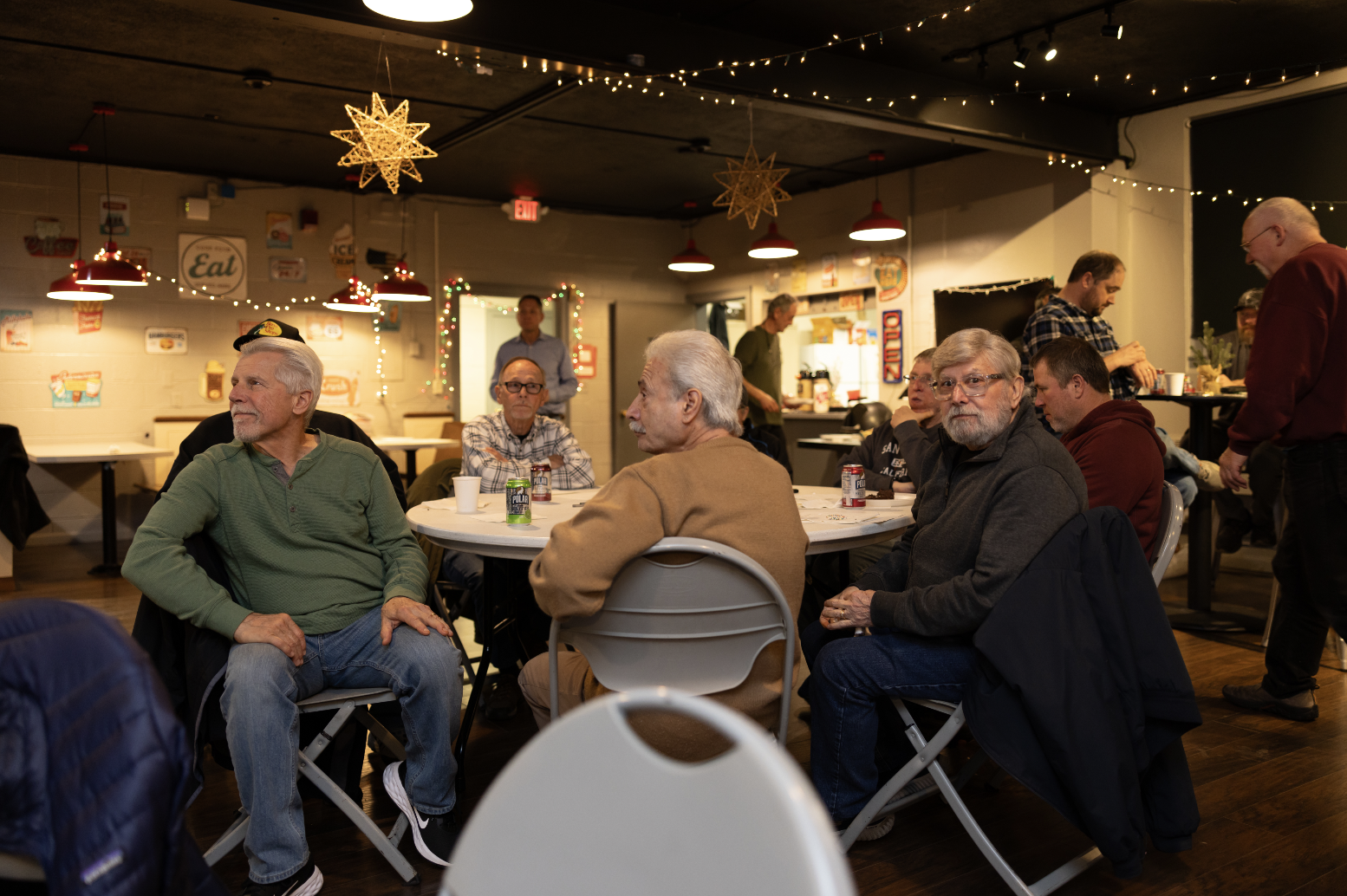 Group of older people sitting around a Christmas-themed decorated table in a cozy restaurant or event space, engaged in conversation and enjoying drinks.