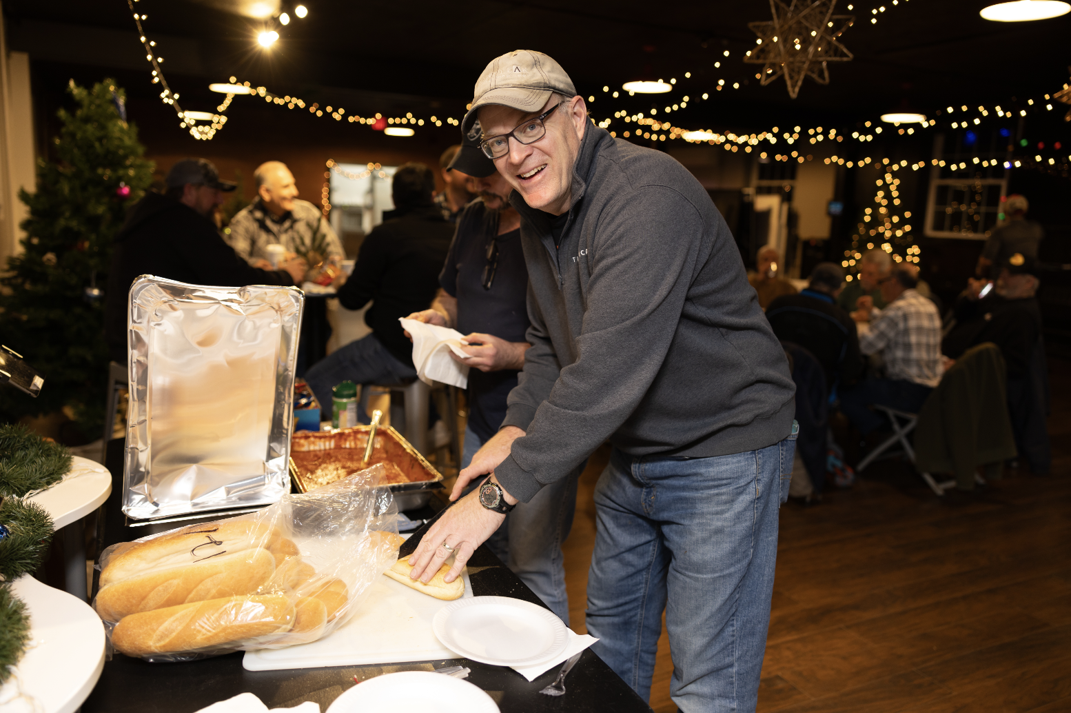 A man with glasses and a gray cap slices bread at a holiday gathering, with a decorated Christmas tree and string lights in the background.