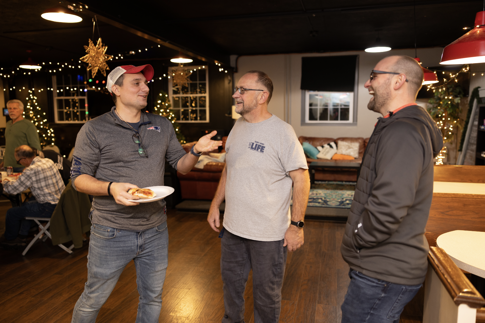 Three men conversing and smiling at a gathering in a decorated indoor space with Christmas lights and trees; one man holding a slice of pizza.