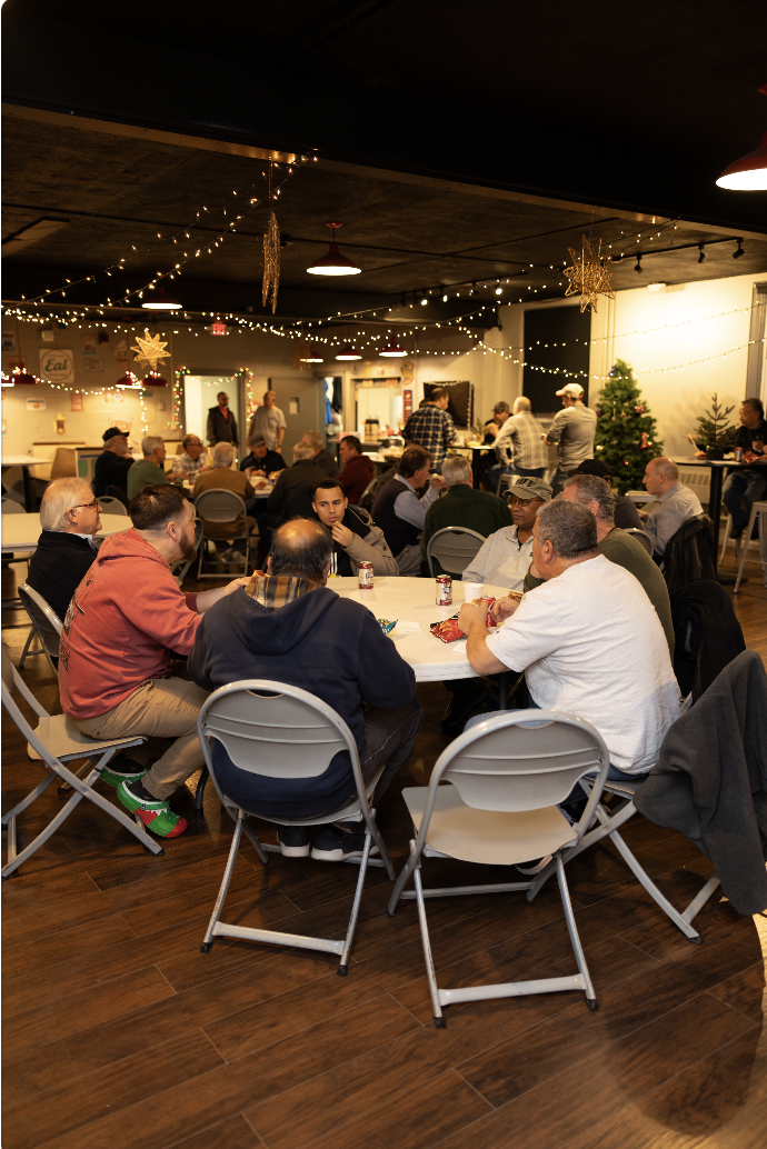 People gathered around tables at a holiday party with festive decorations and a Christmas tree in the background.