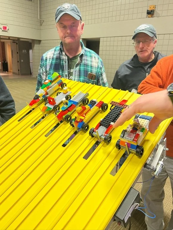 People watching small LEGO cars race down a yellow track with multiple lanes.