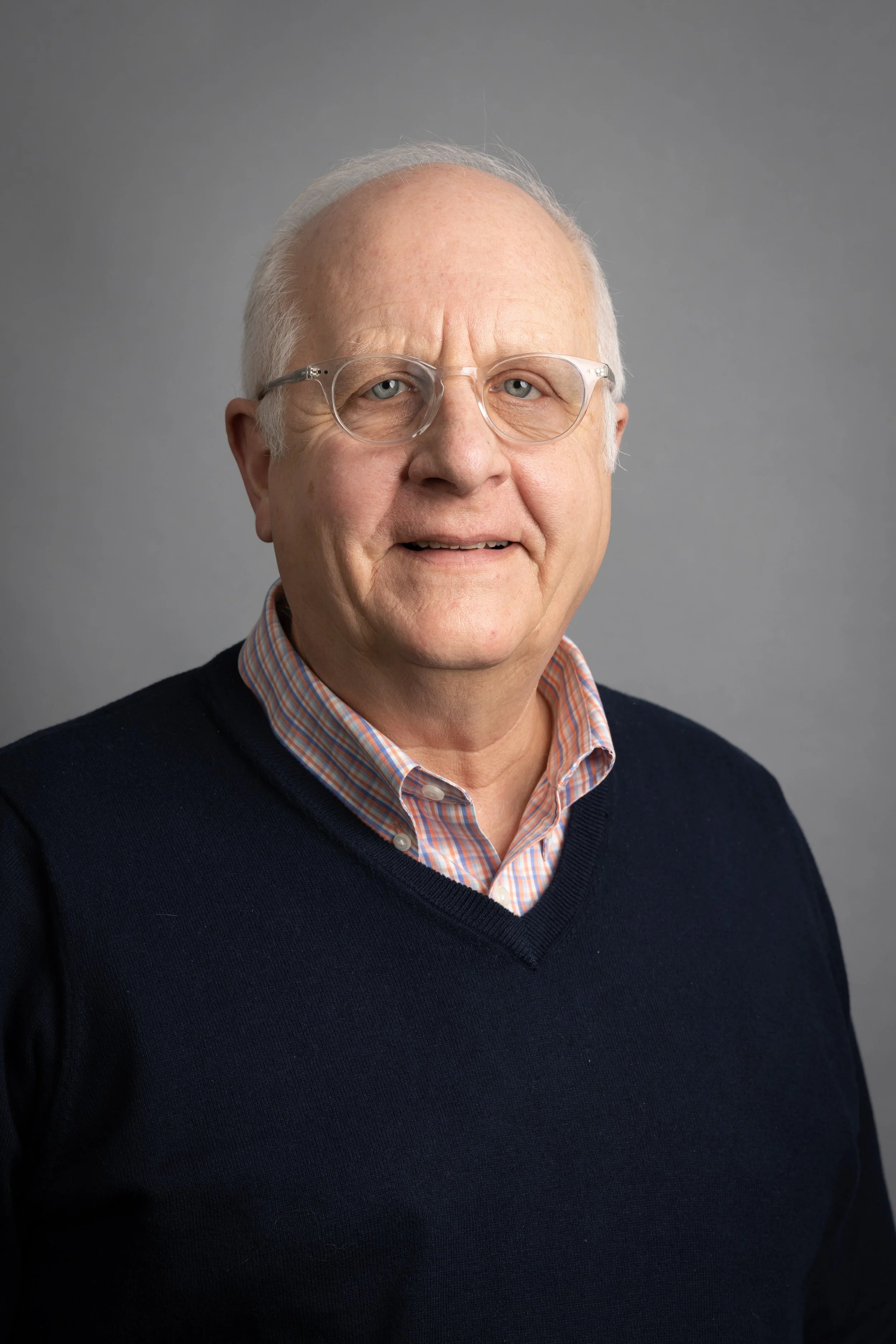 Portrait of an elderly man wearing glasses, a checkered shirt, and a navy sweater against a neutral background.