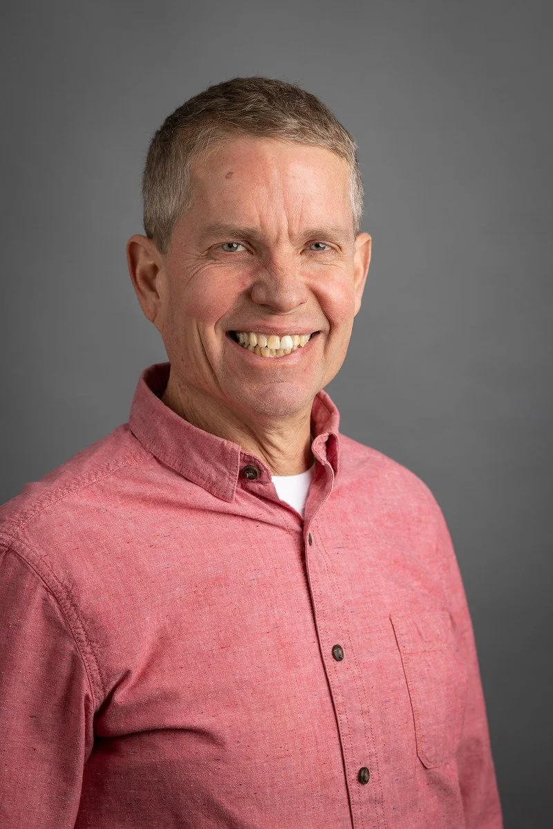 Headshot of a smiling middle-aged man with short light brown hair, wearing a pink button-up shirt against a plain gray background.