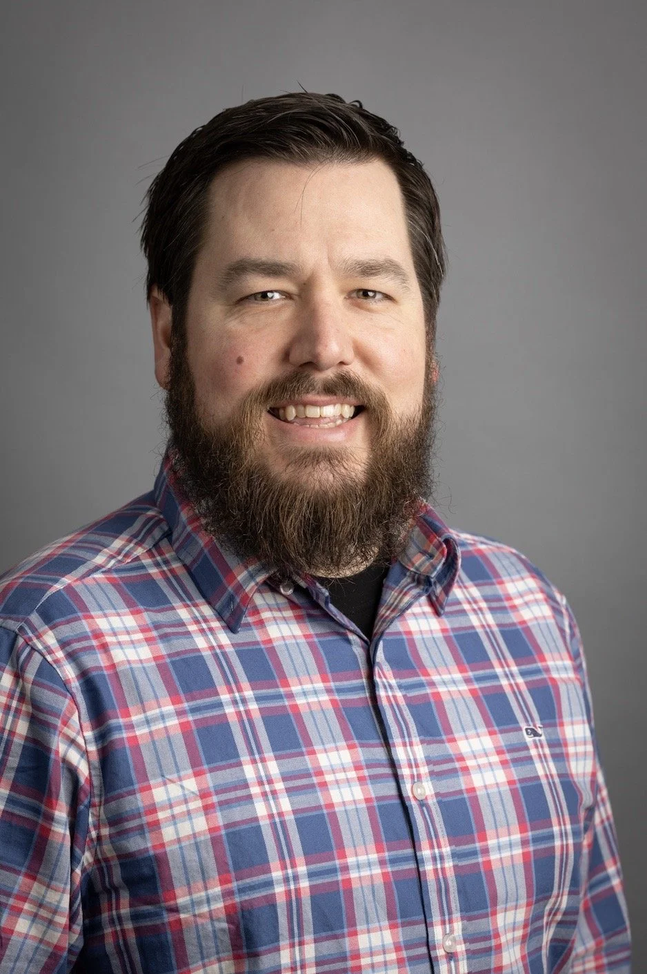 Headshot of man with dark hair, beard, and mustache, wearing a plaid shirt, smiling, against a plain gray background.