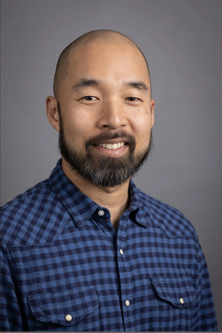 Portrait of a smiling Asian man with a beard, wearing a blue checkered shirt against a gray background.