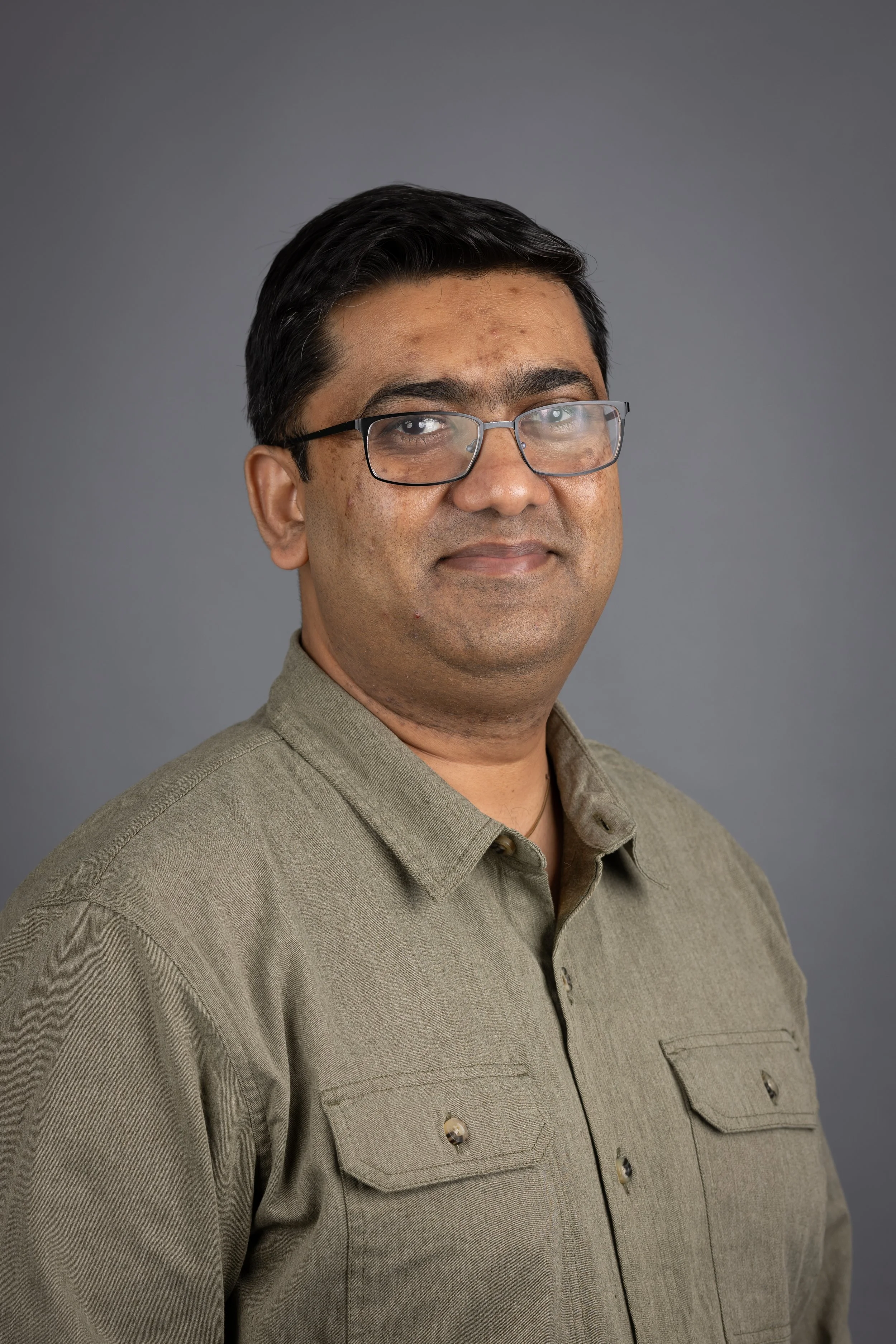 A portrait of a man with glasses, wearing a light brown collared shirt, standing against a gray background.