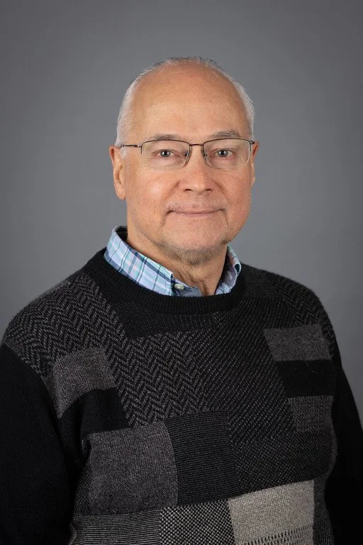 Portrait of an older man with glasses, wearing a plaid shirt and a patterned sweater, standing against a gray background.