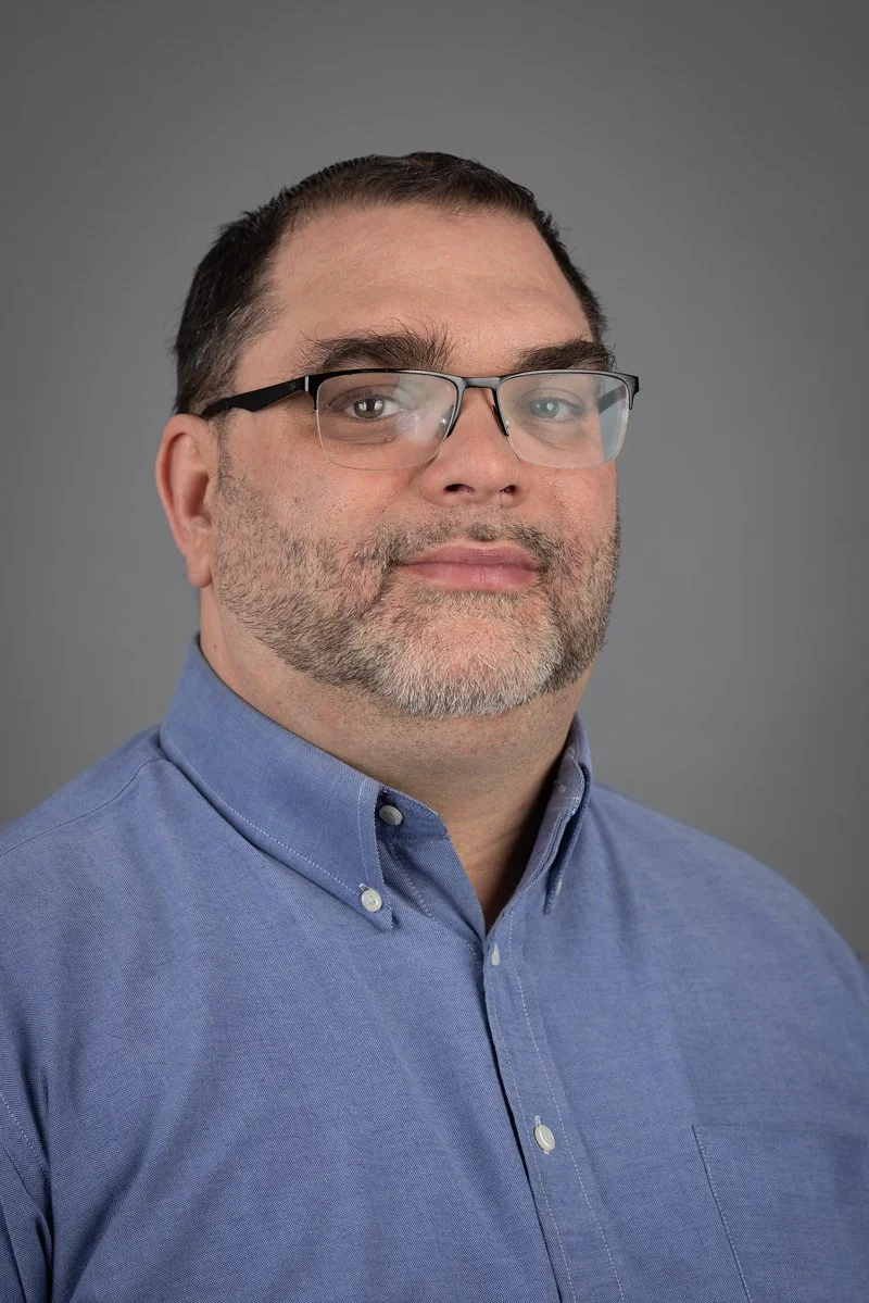 A middle-aged man with dark hair and glasses wearing a blue button-up shirt against a gray background.