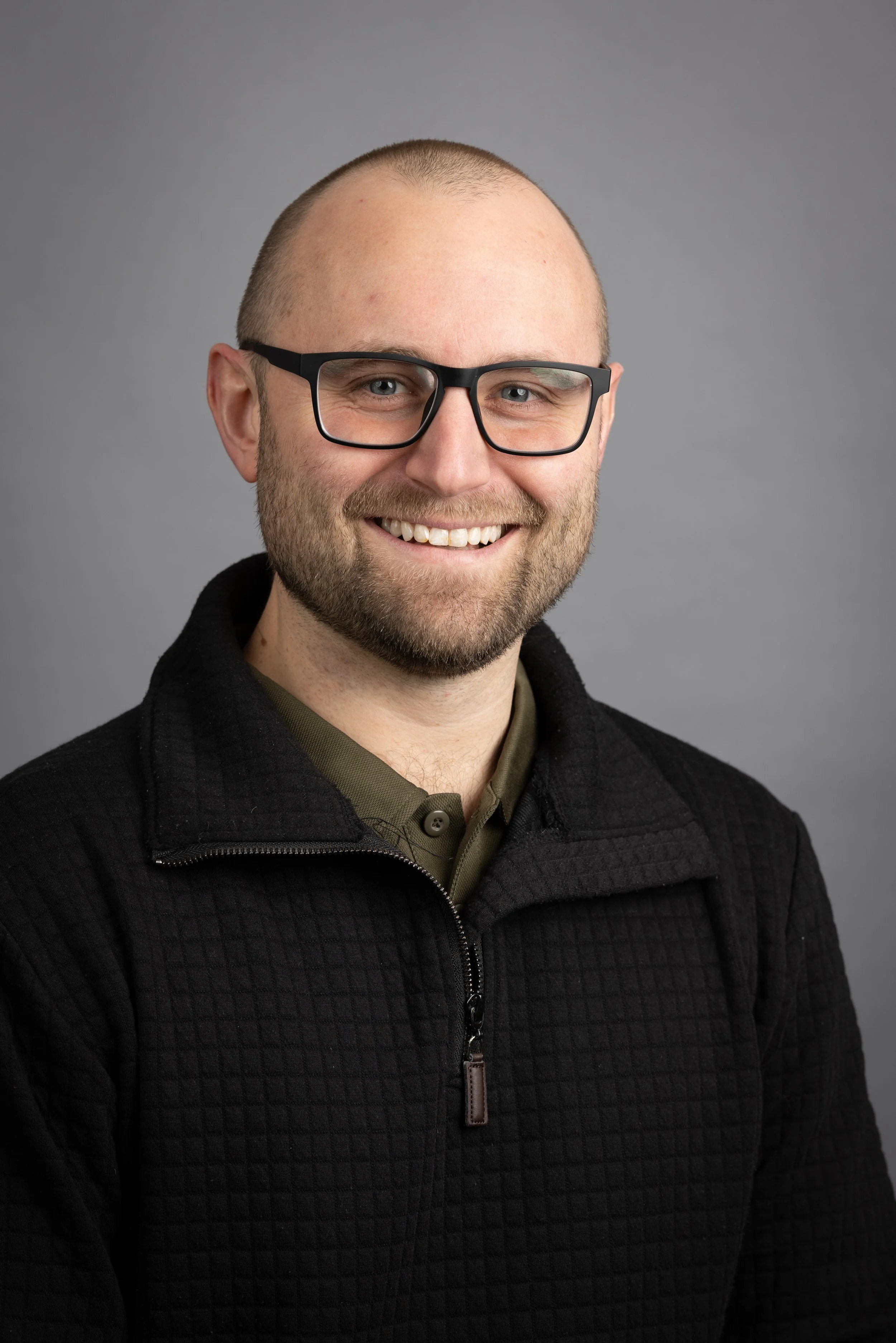 Headshot of a smiling man with glasses, beard, and short hair, wearing a black quilted jacket and a collared shirt, against a gray background.