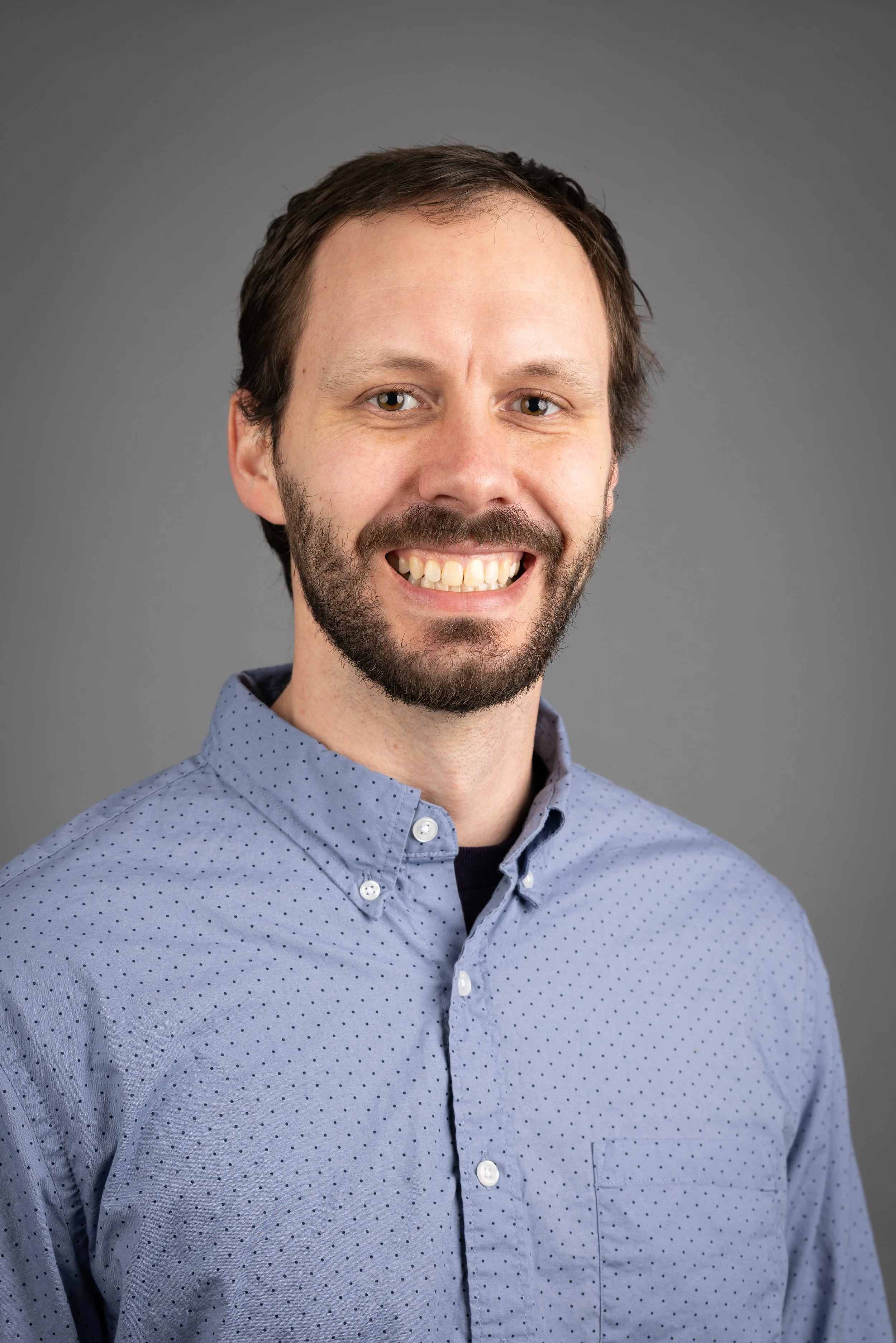 A man with brown hair and beard smiling, wearing a light blue button-up shirt, against a plain gray background.