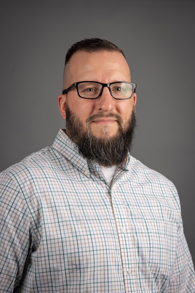 A man with glasses, a trimmed beard, and short hair wearing a checkered button-up shirt, posing against a plain gray background.