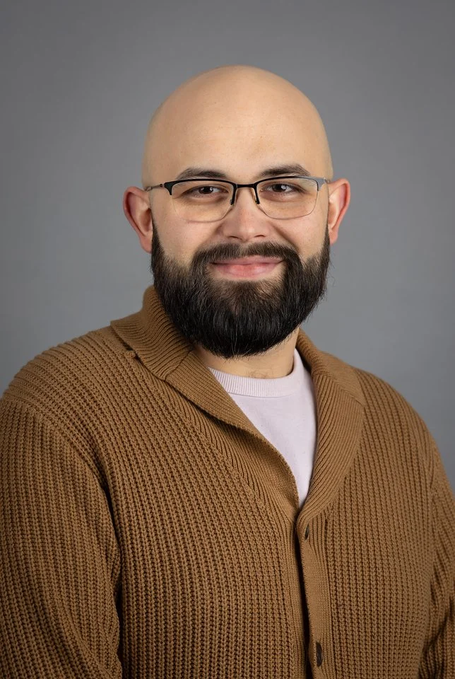 A bald man with a beard wearing glasses and a brown knit sweater, smiling at the camera against a gray background.
