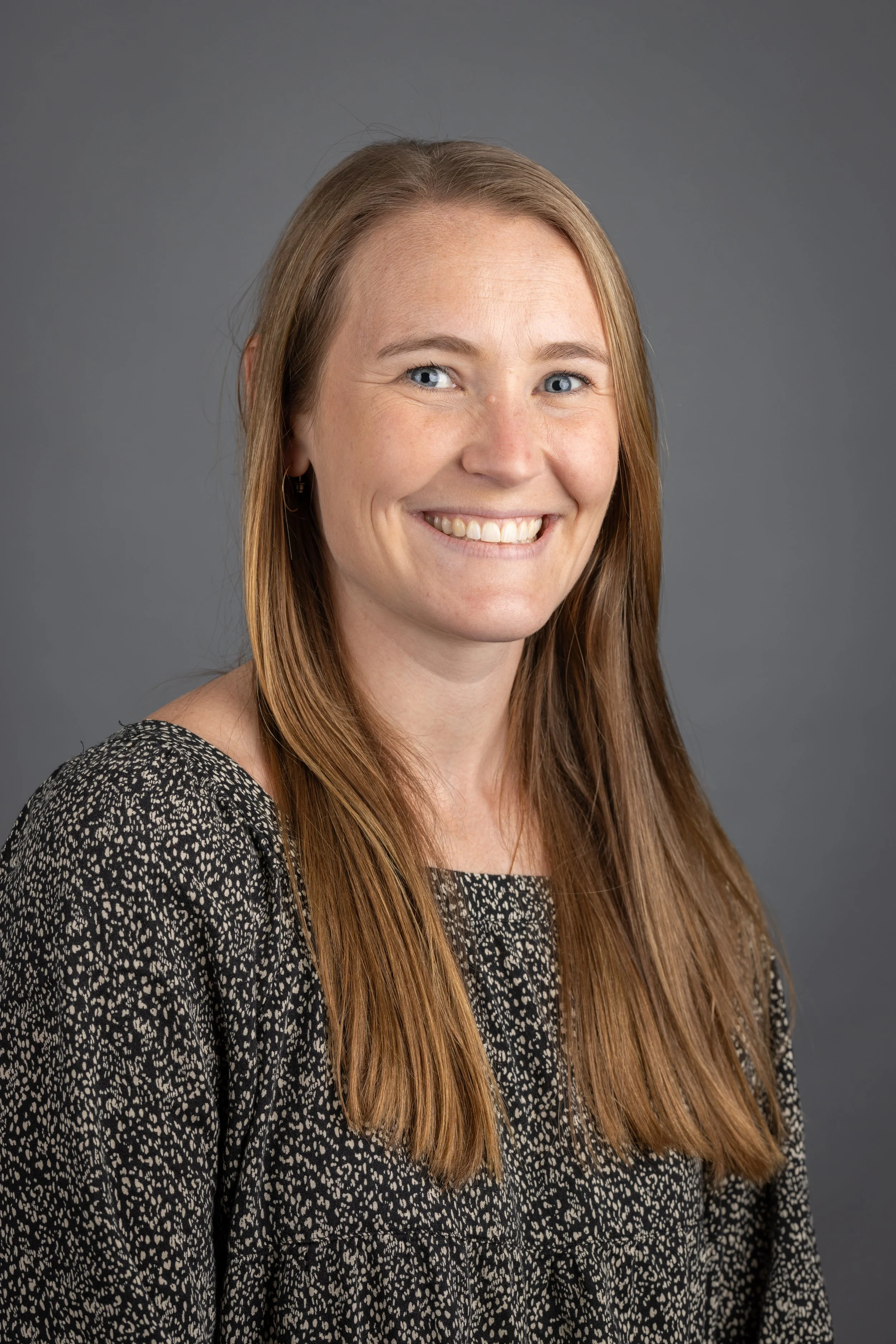 Headshot of a smiling woman with long red hair, wearing a black patterned top, against a grey background.