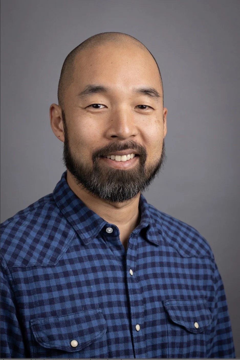 Portrait of a smiling man with a beard, wearing a blue checkered shirt, against a gray background.
