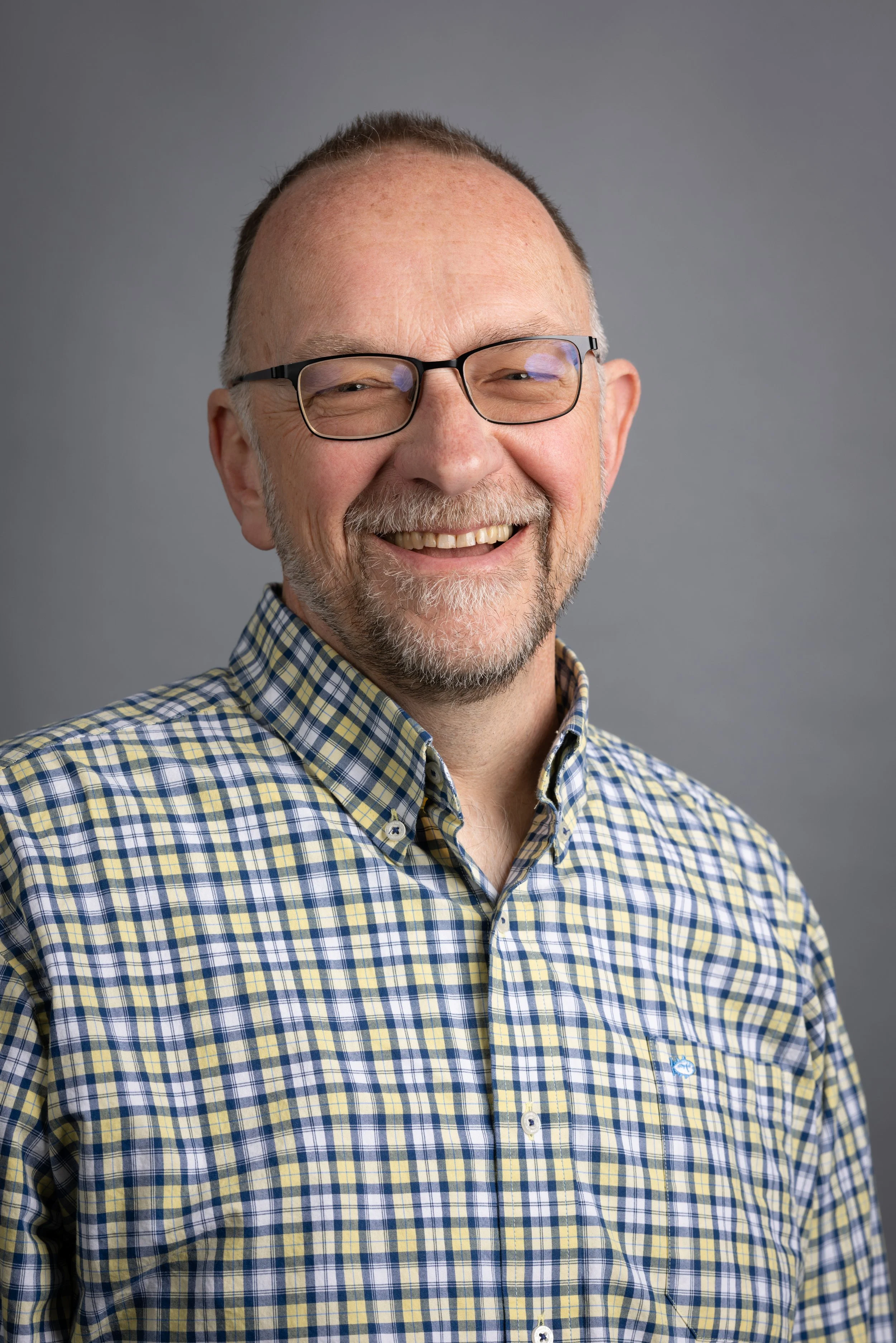 Man wearing glasses and a yellow checkered shirt smiling against a gray background.