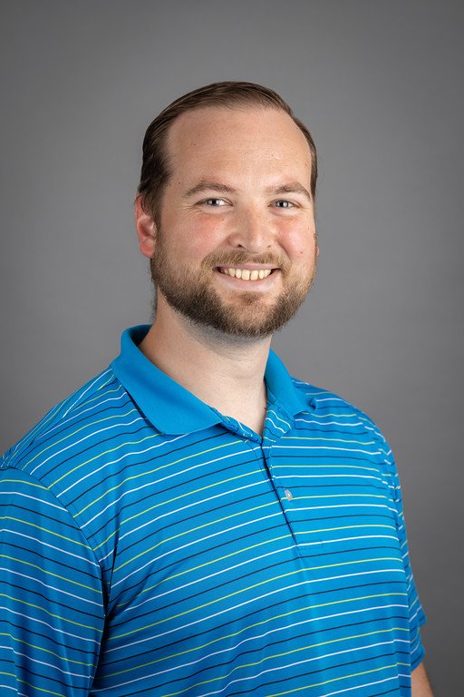 A smiling man with short brown hair and a beard, wearing a blue polo shirt with white and yellow stripes, posing against a gray background.