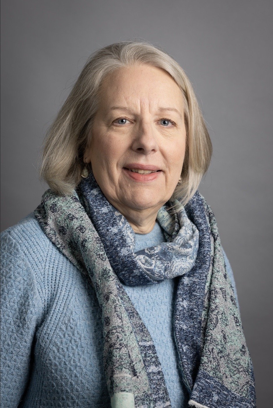 Portrait of a middle-aged woman with shoulder-length gray hair, wearing a light blue sweater and a multicolored scarf, against a plain gray background.