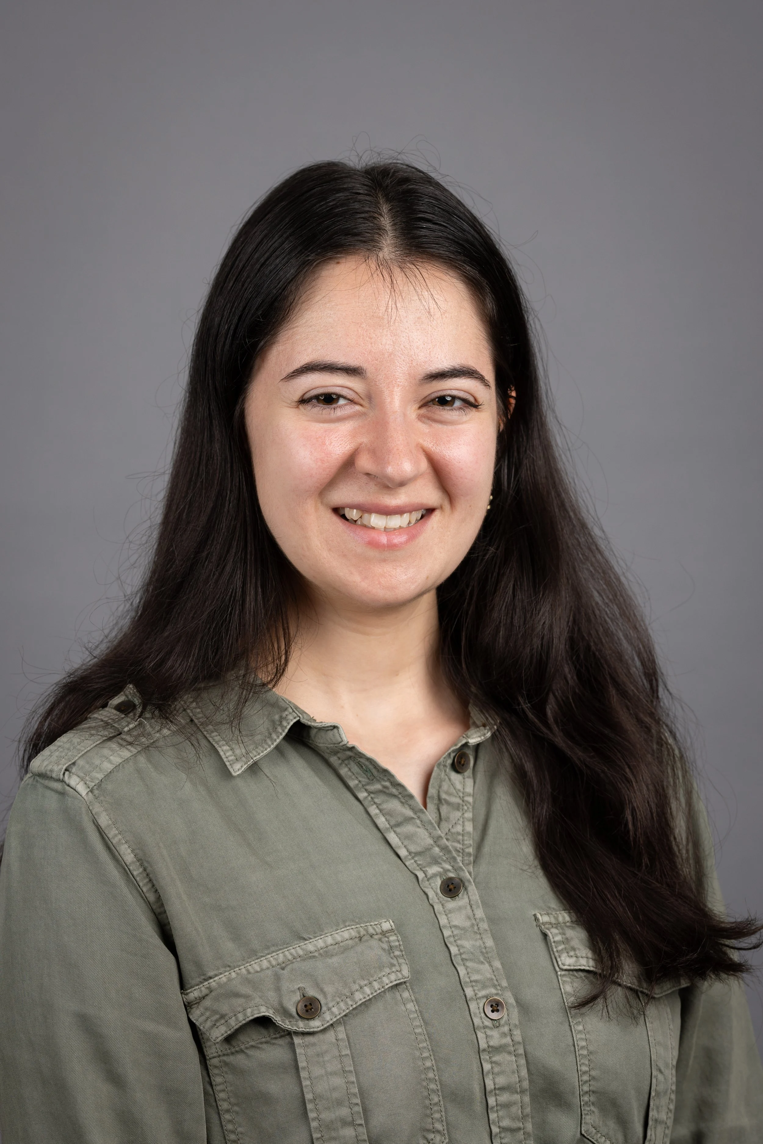 Portrait of a woman with long dark hair wearing an olive green button-up shirt, smiling at the camera against a plain gray background.