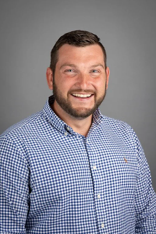 Portrait of a smiling man with short brown hair, a beard, and blue eyes, wearing a blue and white checkered shirt against a neutral gray background.